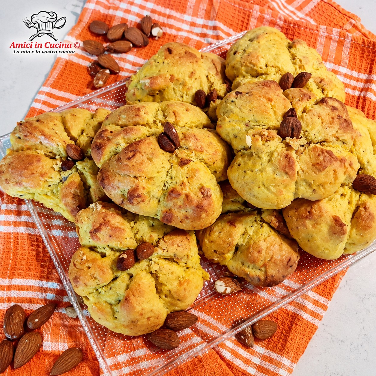Vassoio trasparente colmo di panini salati alla zucca a forma di piccole zucchette, decorati con una mandorla al centro a mo' di picciolo, su un canovaccio a quadri arancioni e bianchi