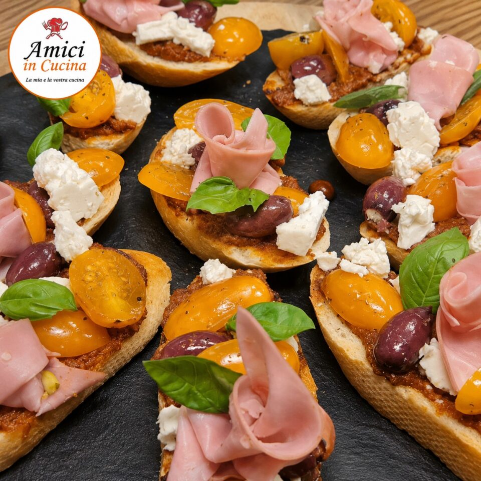 Dettaglio di bruschettine estive croccanti con crema di pomodori secchi, mortadella, feta, pomodorini gialli, olive nere e basilico fresco.