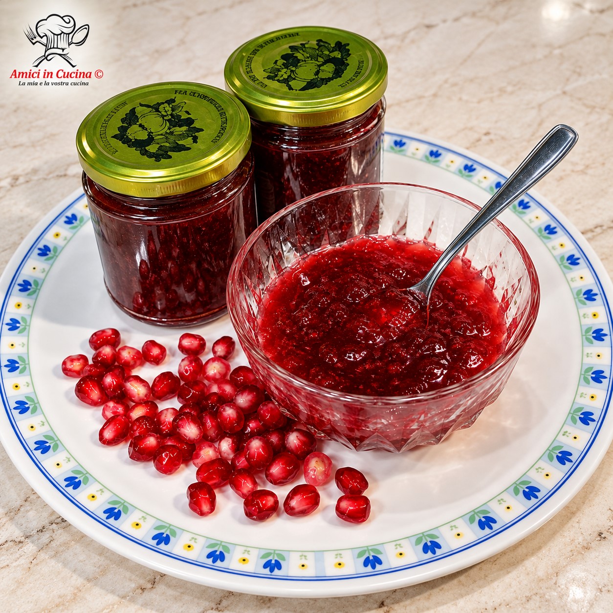 Coppetta di vetro trasparente contenente confettura rossa di melograno con un cucchiaino, affiancata da barattoli pieni e chicchi di melograno sparsi su un piatto bianco decorato