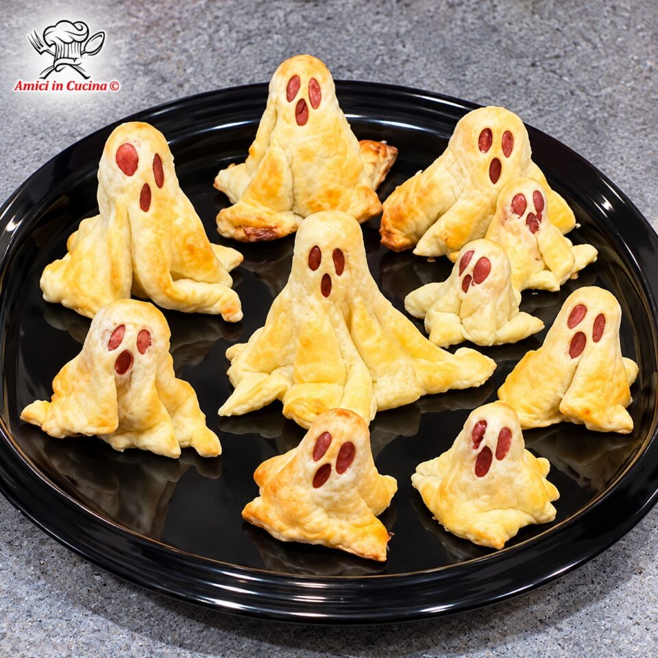 Piccoli rustici a forma di fantasmi realizzati con pasta sfoglia adagiata su wurstel, con fori per occhi e bocca, disposti su un vassoio scuro decorato con lucine