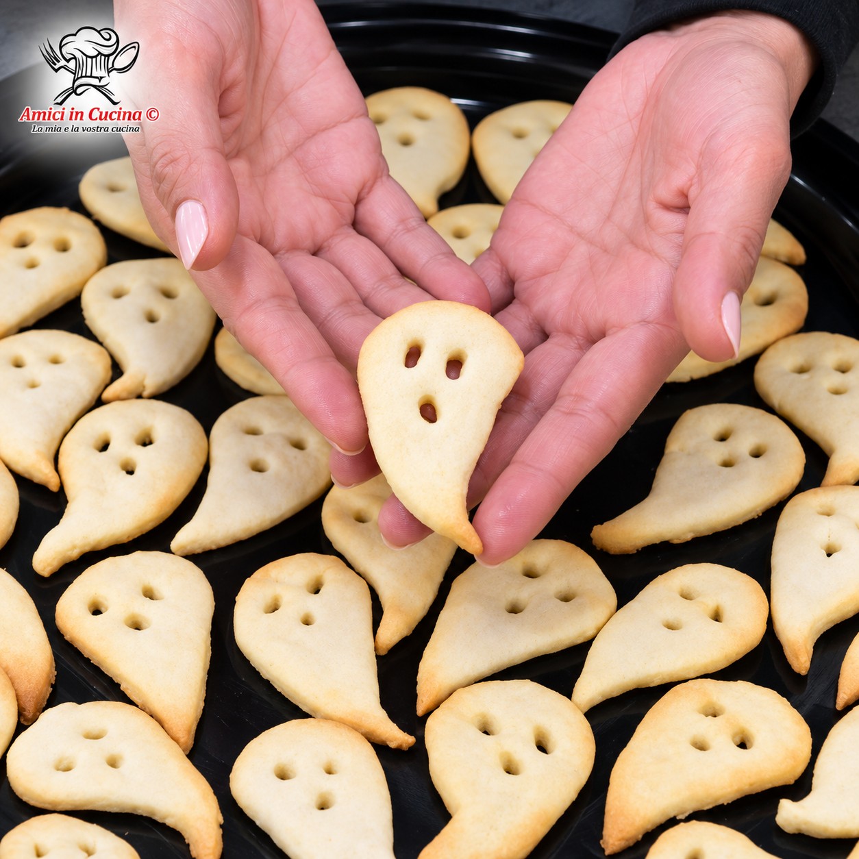 Mani femminili che mostrano un biscotto a forma di fantasmino con tre fori per occhi e bocca, con un vassoio nero pieno di altri biscotti simili sullo sfondo