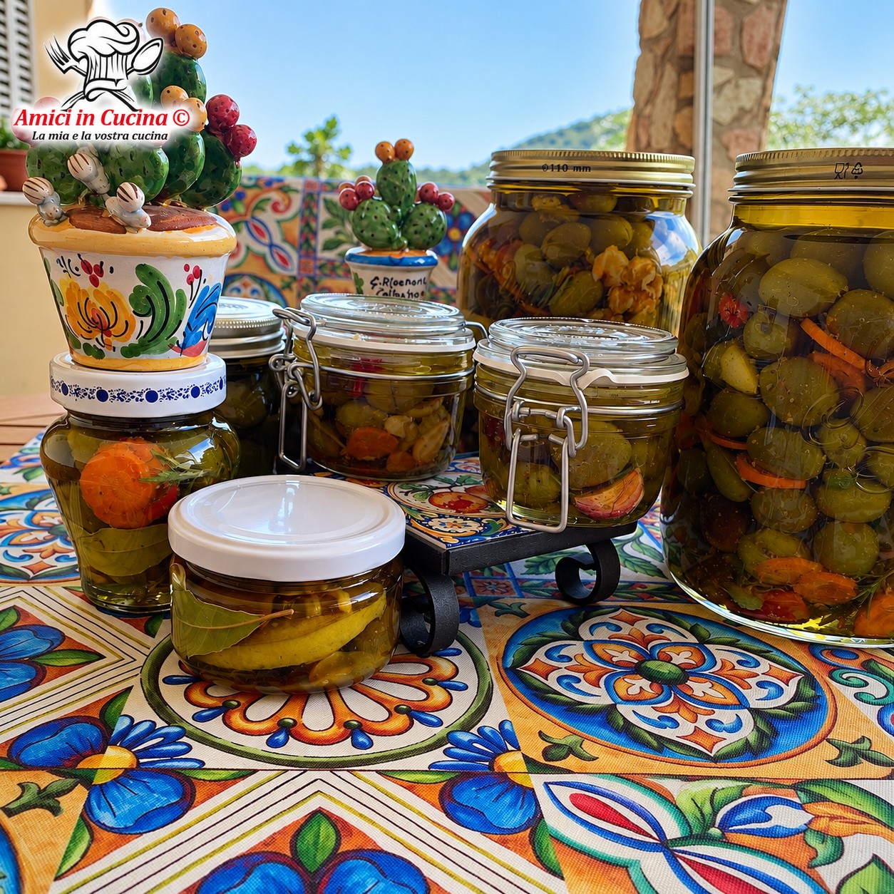 Composizione di barattoli di vetro di diverse dimensioni, pieni di olive verdi schiacciate sott'olio e aromi, adagiati su un piano decorato con ceramiche dai motivi floreali mediterranei