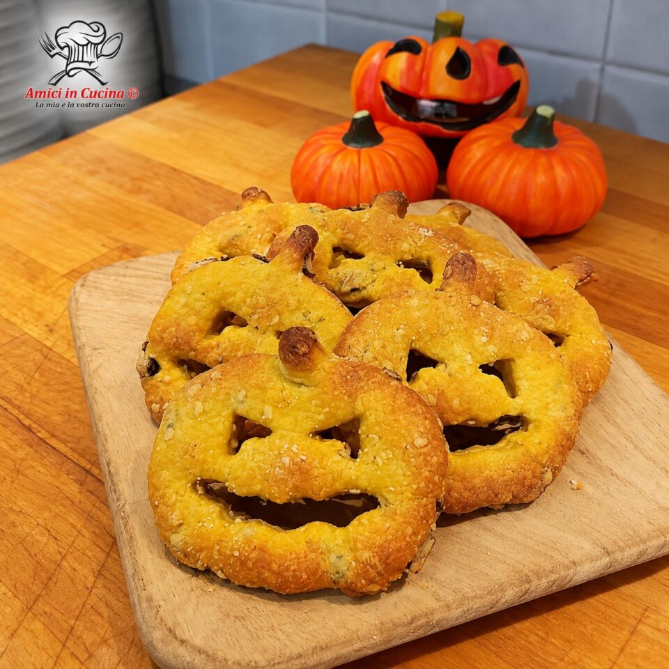 Piccoli rustici salati a forma di zucca intagliata, dal colore dorato, disposti su un tagliere di legno con piccole zucche decorative sullo sfondo