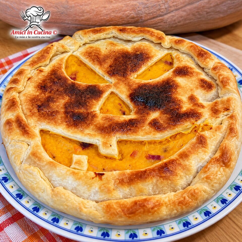 Torta salata di pasta sfoglia farcita con zucca e speck, con la superficie intagliata a forma di faccia spaventosa di zucca di Halloween, servita su un piatto bianco con decori blu