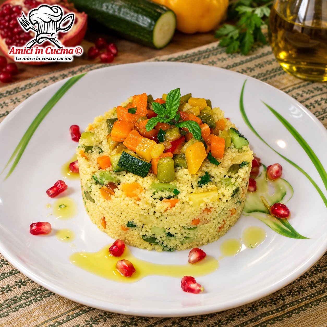 Piatto bianco elegante con un tortino rotondo e compatto di cous cous misto a cubetti di verdure croccanti, guarnito in cima con altre verdure, foglie di menta fresca e circondato da chicchi rossi di melograno