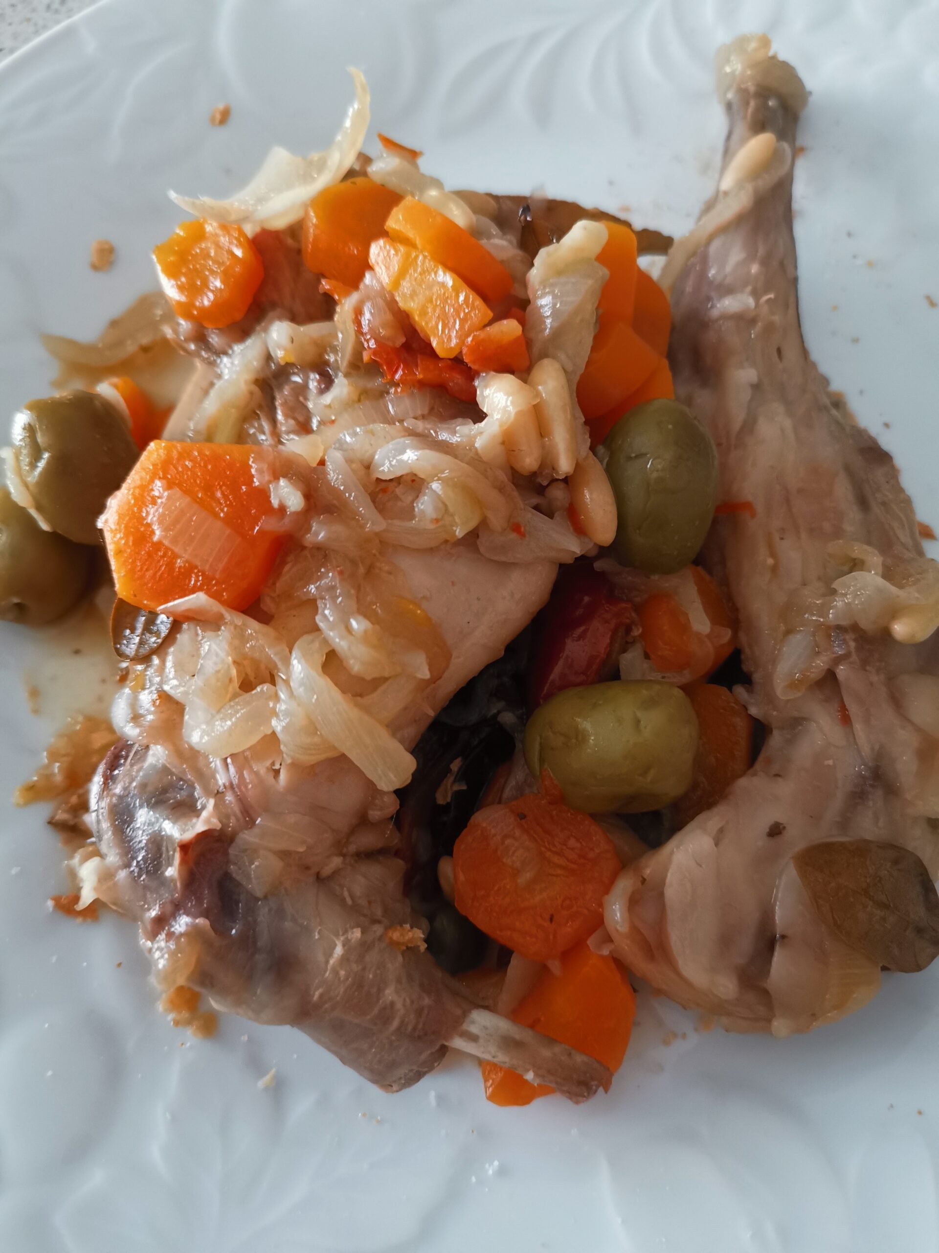 Un primo piano ravvicinato di una porzione di coniglio in umido servito su un piatto bianco, condito con carote a rondelle, olive verdi, pomodori secchi, cipolla stufata e pinoli tostati.
