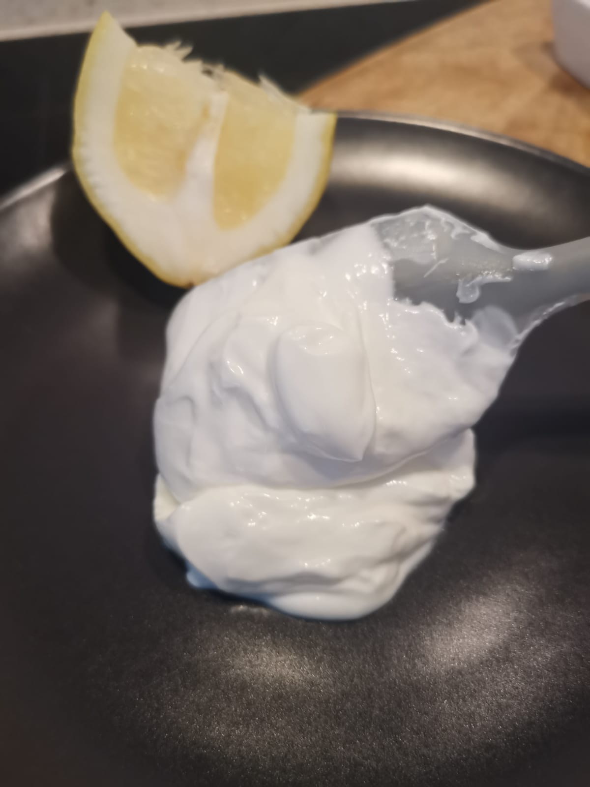 Un piatto nero con una cucchiaiata densa e cremosa di maionese allo skyr senza uova accanto a uno spicchio di limone
