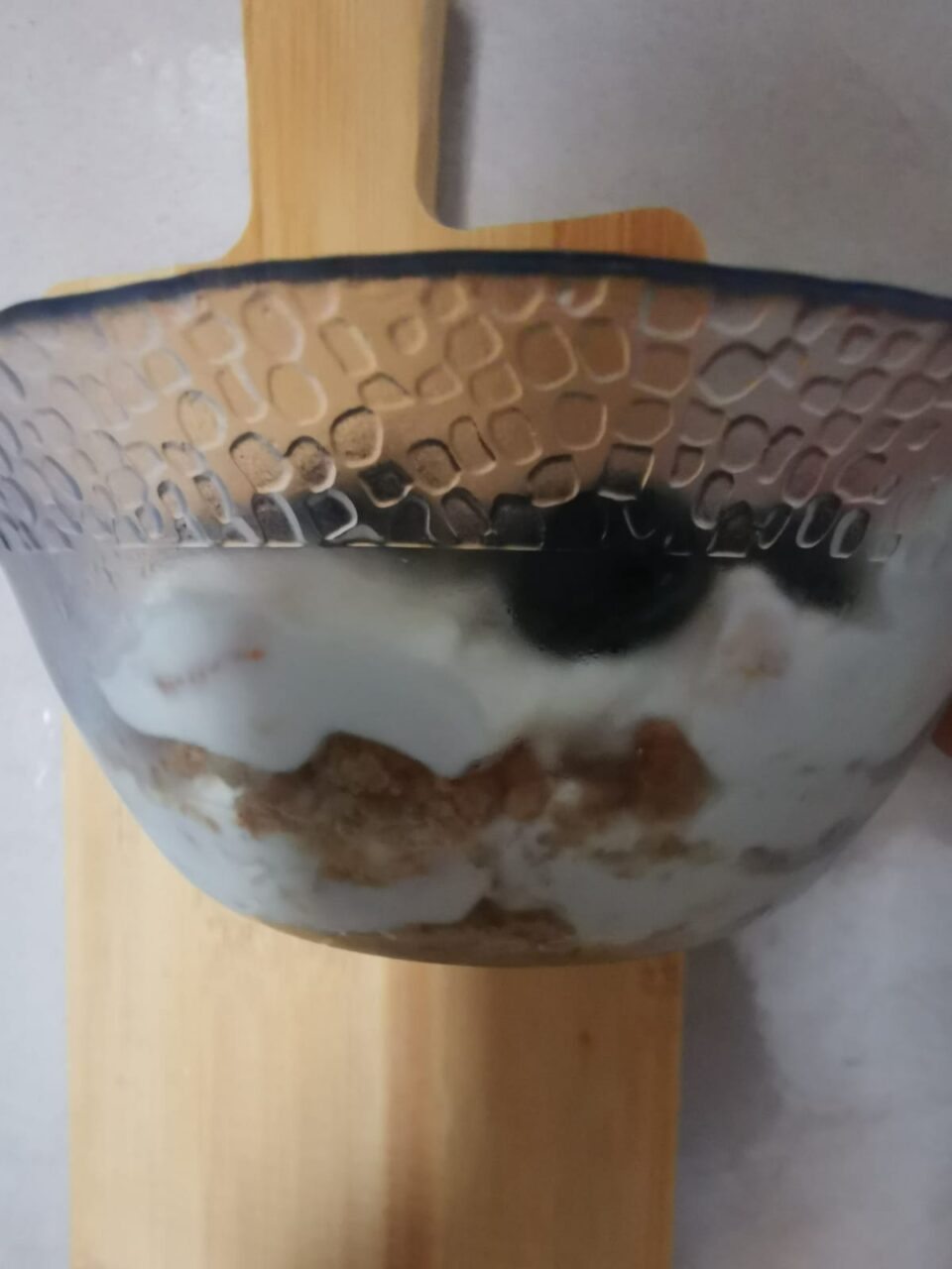 Una coppetta di vetro trasparente con un dolce alla ricotta veloce, pesche nettarine e mirtilli, servita su un tagliere di legno chiaro.