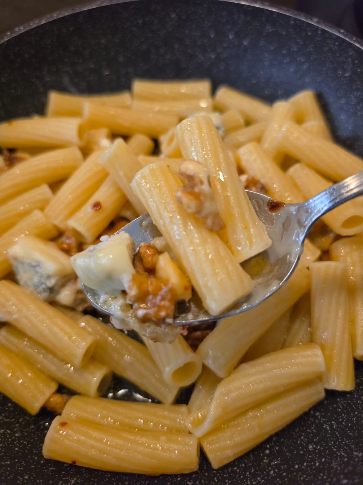 Primo piano di un cucchiaio che solleva rigatoni conditi con gorgonzola fuso, noci e peperoncino, sopra la padella.