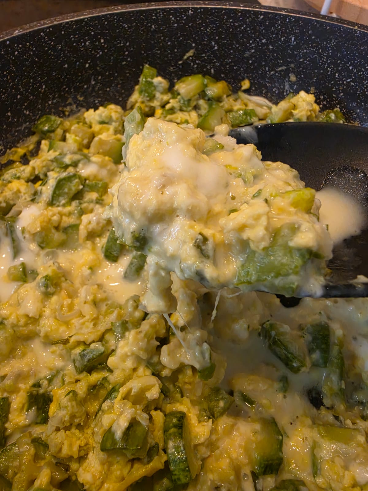 Mestolo che solleva una porzione di zucchine uova e stracchino in padella con formaggio filante e cremoso.