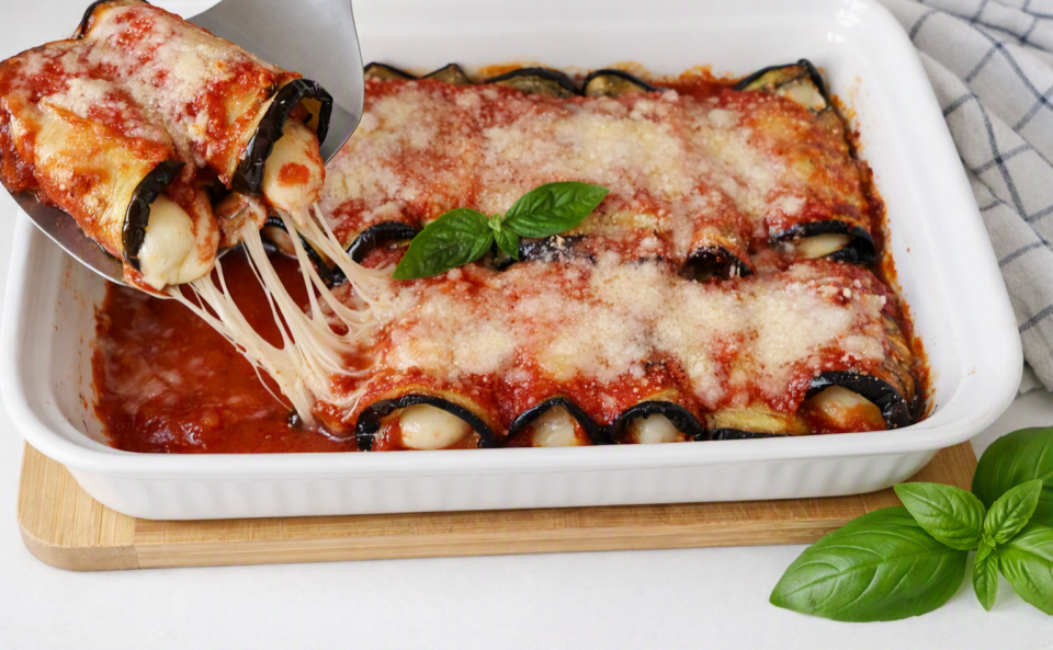 Immagine di una teglia di involtini di melanzane leggeri e filanti, disposti ordinatamente in una pirofila bianca. Gli involtini sono ricoperti da sugo di pomodoro e da una gratinatura dorata di Parmigiano che mette in risalto il formaggio fuso. 
