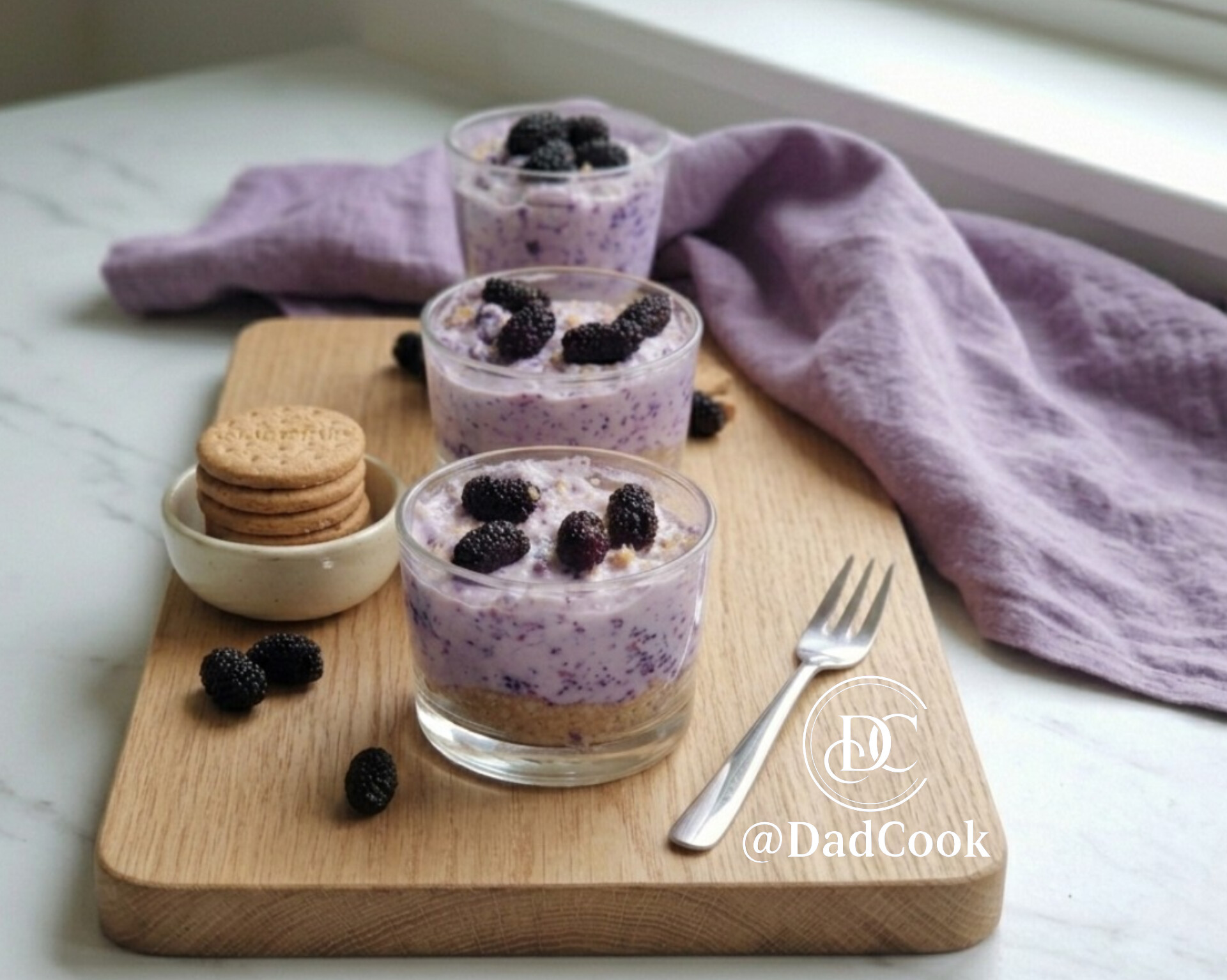 Tre bicchieri di dessert monoporzione con base di biscotto Digestive sbriciolato, crema variegata alla stracciatella, ricotta e gelsi neri, decorati in superficie con gelsi interi, disposti su un tagliere di legno accanto a una pila di biscotti e un canovaccio lilla.