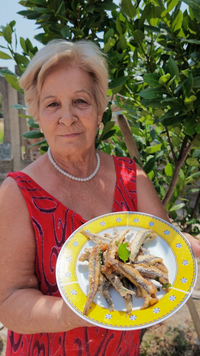 Mamma sorridente all'aperto che mostra un piatto di sarde fritte dorate condite con aceto, peperoncino e menta