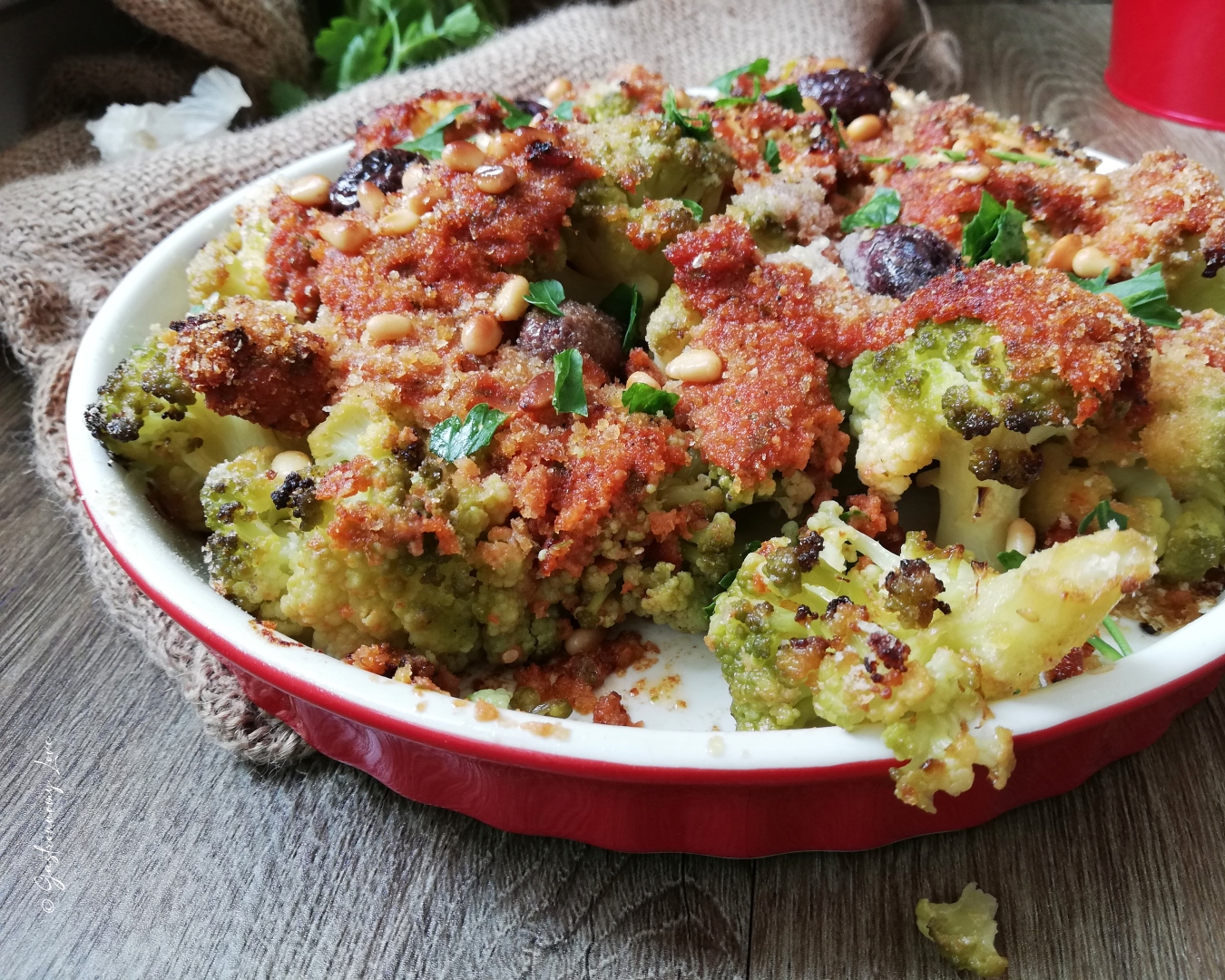 Contorno di cavolfiore verde gratinato al forno con crema di pomodoro, olive nere e pinoli tostati, servito in una teglia rustica rossa.
