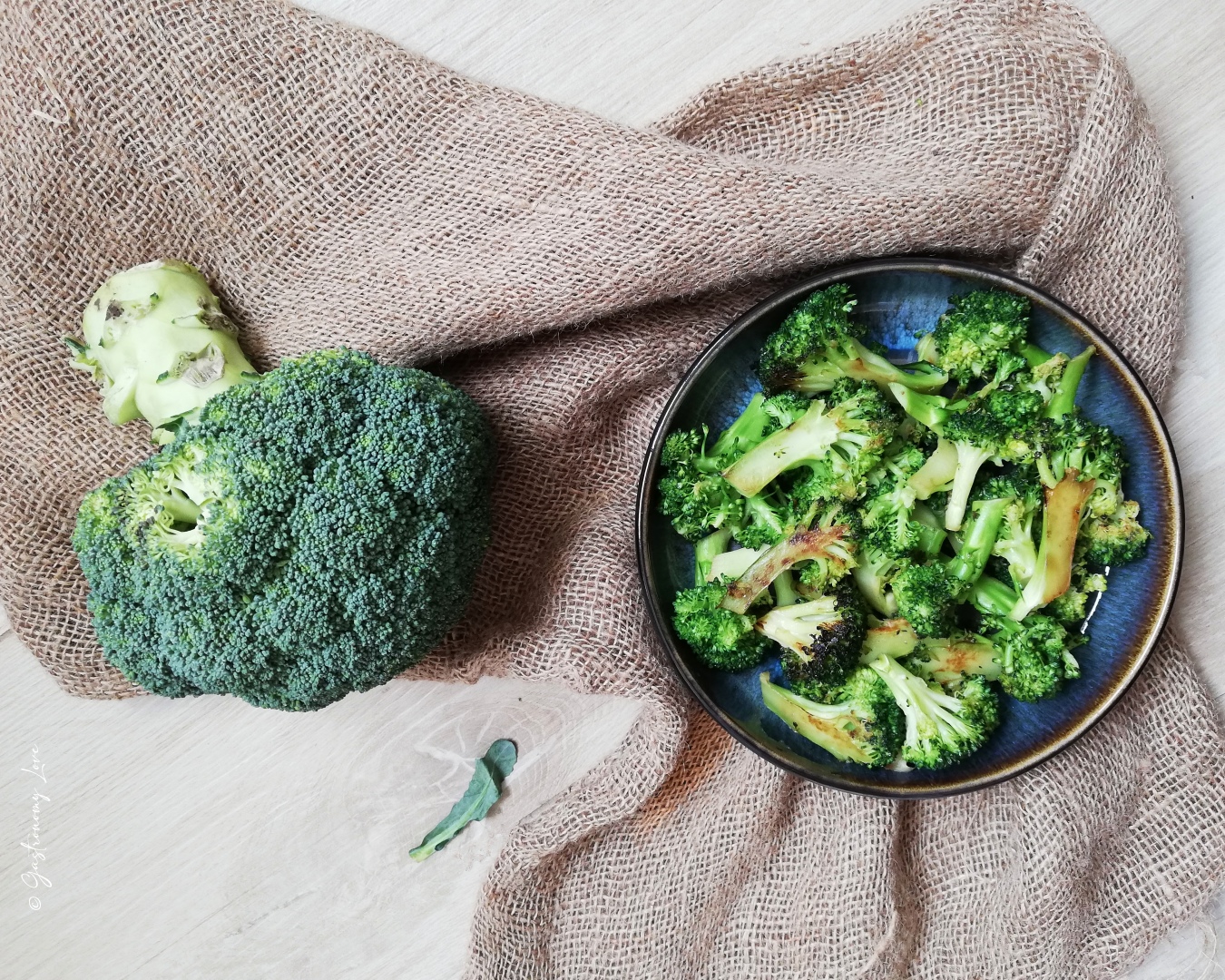 Broccoli arrostiti in padella in un piatto blu poggiato su della iuta e con accanto un broccolo intero crudo