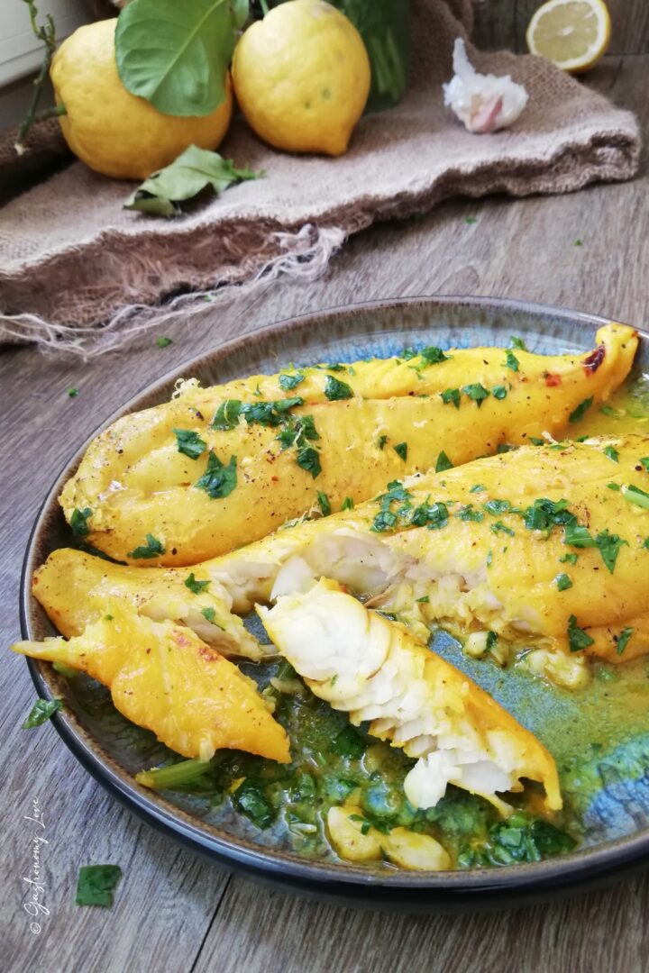 Filetti di merluzzo allo zafferano e limone con un meraviglioso color oro. Sono serviti in un piatto blu e nero e decorati con prezzemolo fresco. Nello sfondo ci sono limoni poggiati su un telo di iuta.