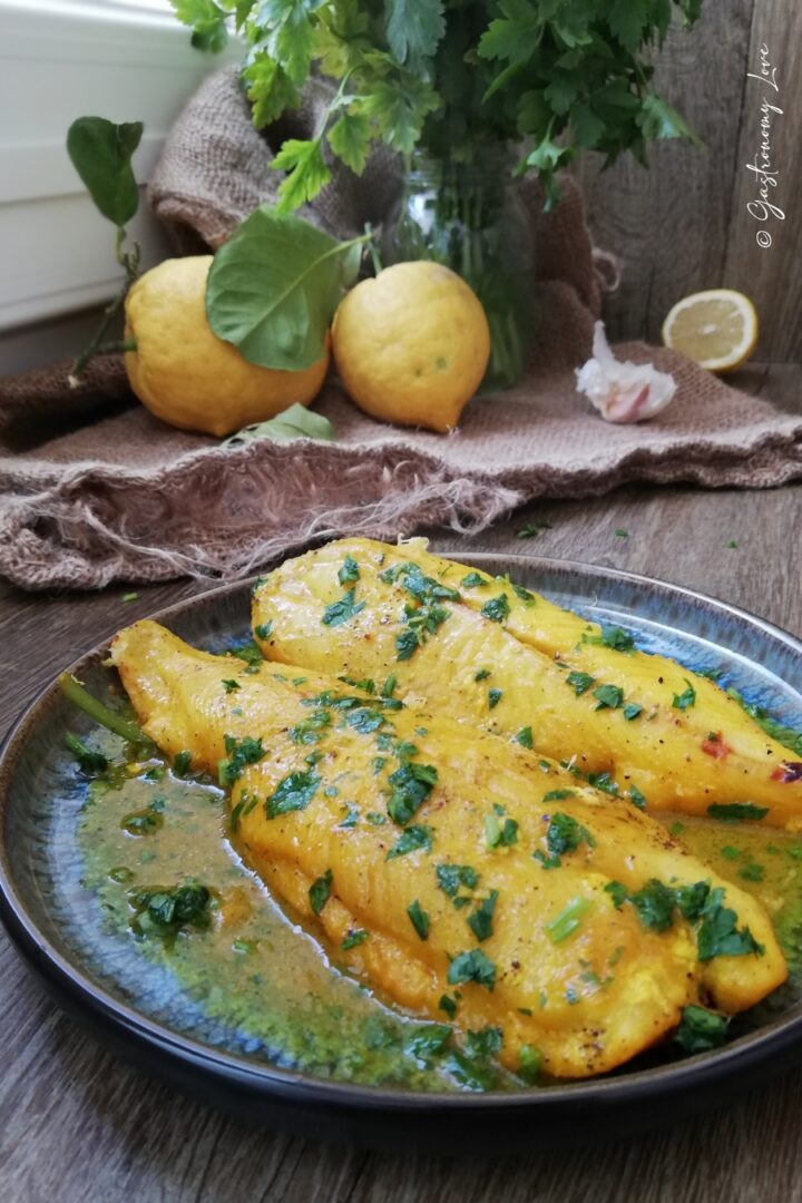 Filetti di merluzzo allo zafferano e limone con un meraviglioso color oro. Sono serviti in un piatto blu e nero e decorati con prezzemolo fresco. Nello sfondo ci sono limoni poggiati su un telo di iuta.