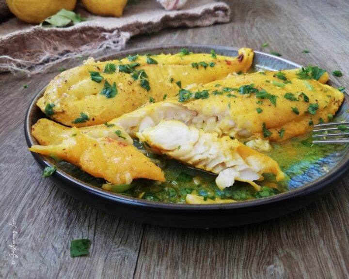 Ci sono dei filetti di merluzzo allo zafferano e limone con un meraviglioso color oro. Sono serviti in un piatto blu e nero e decorati con prezzemolo fresco. Nello sfondo ci sono limoni poggiati su un telo di iuta.