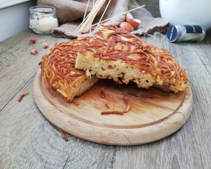 Frittata di pasta alla napoletana alta e dorata su un tagliere di legno, con spaghetti croccanti in superficie, uova, basilico fresco e ingredienti rustici sullo sfondo.