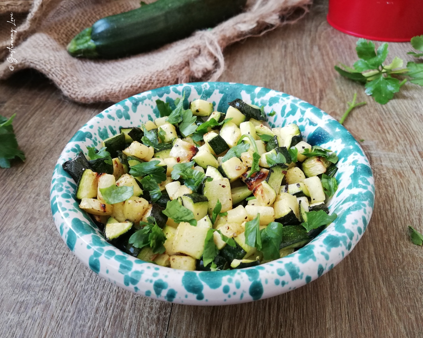 Primo piano di una ciotola rustica bianca e verde acqua piena di zucchine a cubetti cucinate nella friggitrice ad aria, condite con prezzemolo fresco e dorate alla perfezione.