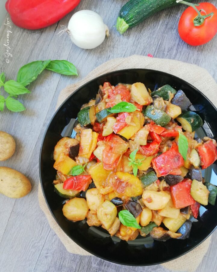 Vista dall'alto di un piatto nero colmo di ciambotta napoletana posizionato su un tavolo di legno grigio, circondato da ingredienti crudi come patate, cipolla, zucchina e pomodoro.