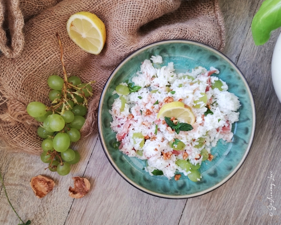Foto orizzontale di un'insalata di riso estiva con prosciutto crudo a listarelle, acini di uva bianca e gherigli di noci, servita in una ciotola di ceramica rustica su un tavolo di legno. La composizione include anche un grappolo d'uva fresca e mezzo limone.