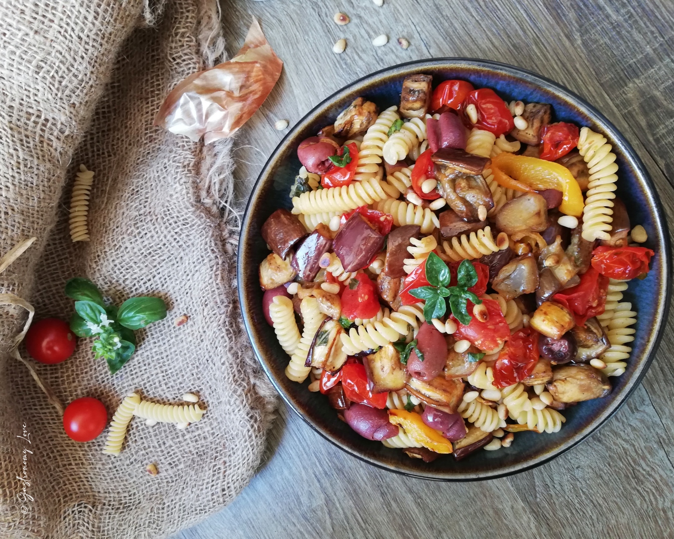Inquadratura dall'alto di un piatto rustico di pasta all'ortolana con fusilli, pomodorini, melanzane, peperoni, olive e pinoli tostati.