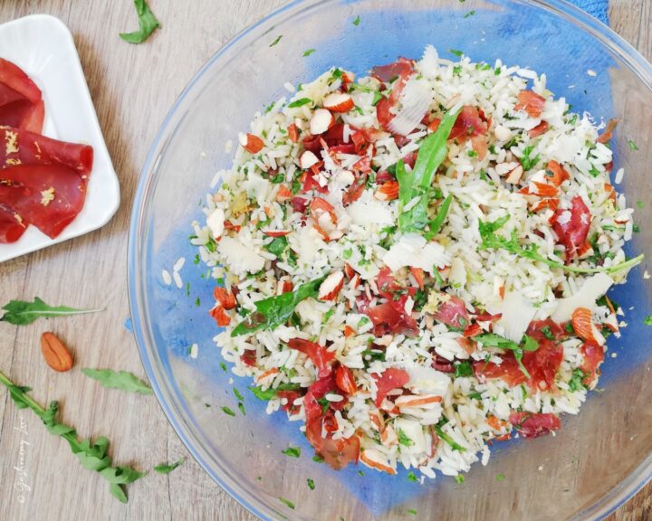 Inquadratura orizzontale di un'insalata di riso sfiziosa con bresaola, rucola tagliata finemente, scaglie di grana e mandorle in una ciotola di vetro, con ingredienti freschi sul tavolo di legno. Ricetta estiva veloce di Gastronomy Love.