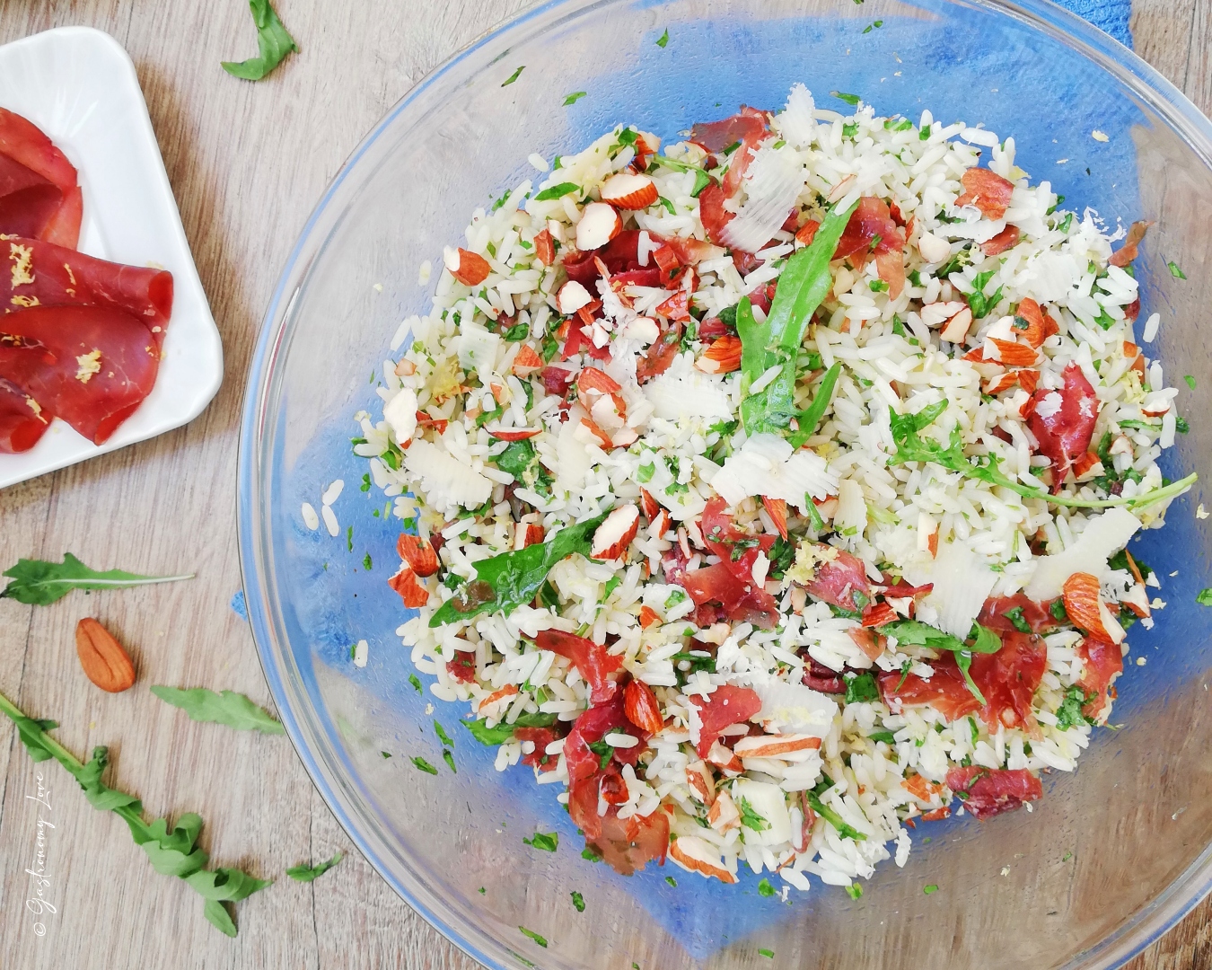 Inquadratura orizzontale di un'insalata di riso sfiziosa con bresaola, rucola tagliata finemente, scaglie di grana e mandorle in una ciotola di vetro, con ingredienti freschi sul tavolo di legno. Ricetta estiva veloce di Gastronomy Love.