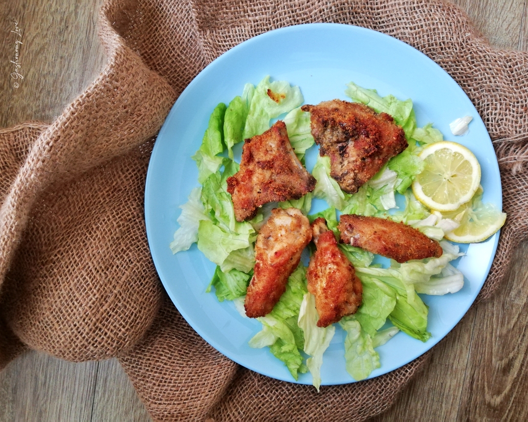 Veduta dall'alto di un piatto azzurro con alette e stuzzichini di pollo panati e dorati, adagiati su un letto di lattuga con fette di limone. Il piatto è appoggiato su una superficie in legno accanto a un tessuto di juta marrone arricciato. In alto a sinistra è presente il watermark "© Gastronomy Love".