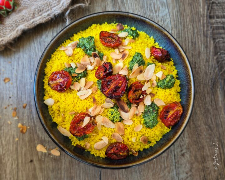Inquadratura zenitale dall'alto di un piatto fondo scuro riempito di couscous alla curcuma color oro. La ricetta è guarnita con pesto di basilico, pomodorini caramellati e mandorle a lamelle. Lo sfondo mostra un tavolo in legno rustico con briciole e scaglie di mandorle sparse attorno al piatto.