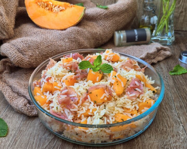 Insalata di riso prosciutto e melone servita in una ciotola di vetro su un tavolo di legno, con una fetta di melone cantalupo e un vasetto di pepe nero sullo sfondo.