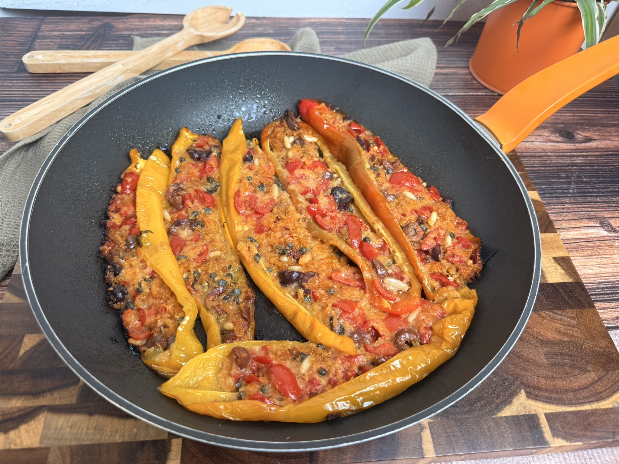 Peperoni ripieni in padella senza forno: facili, veloci e saporiti, ripieni di mollica di pane, pomodorini, parmigiano grattugiato, capperi, pinoli e basilico. Disposti su un piatto bianco. Il ripieno è dorato e leggermente gratinato, decorati con foglie di basilico fresco