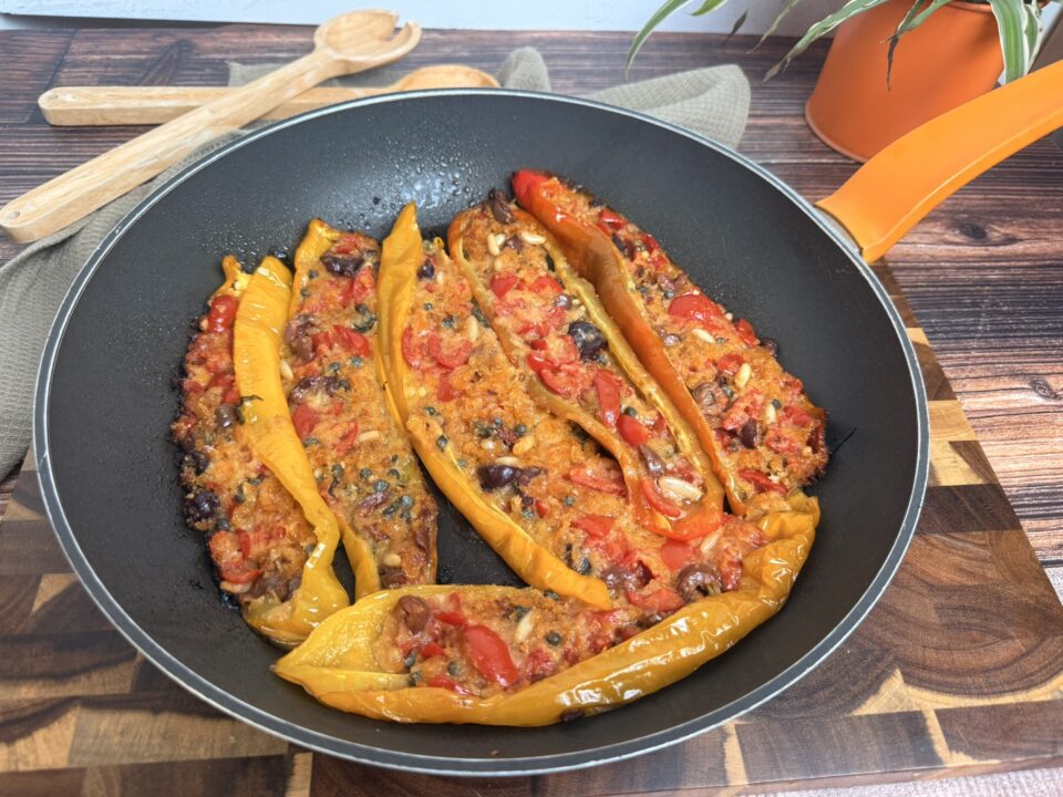 Peperoni ripieni in padella senza forno: facili, veloci e saporiti, ripieni di mollica di pane, pomodorini, parmigiano grattugiato, capperi, pinoli e basilico. Disposti su un piatto bianco. Il ripieno è dorato e leggermente gratinato, decorati con foglie di basilico fresco