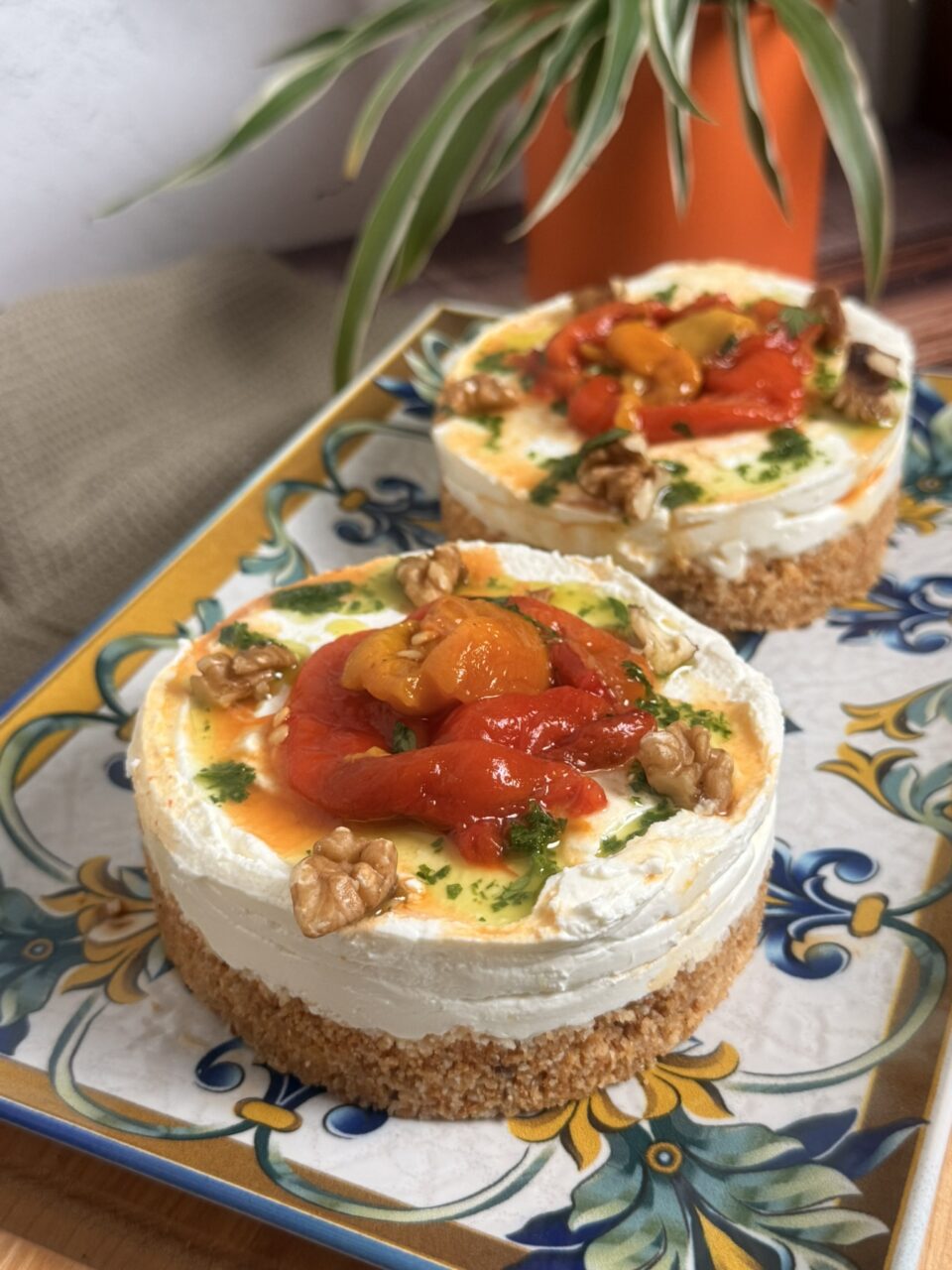 Mini cheesecake salate senza cottura: l’antipasto estivo facile e cremoso con base croccante di taralli, crema di formaggio spalmabile e robiola, topper di peperoni arrostiti, gherigli di noce e olio aromatizzato al prezzemolo, servite su un piatto bianco.