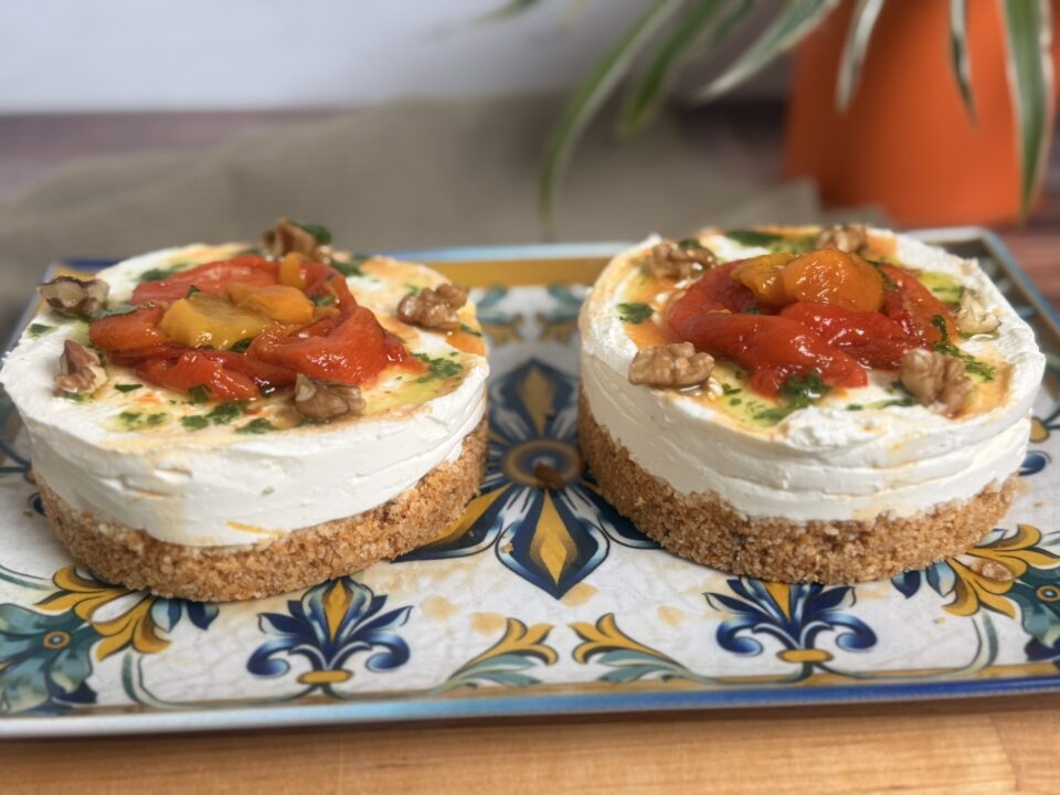 Mini cheesecake salate senza cottura: l’antipasto estivo facile e cremoso con base croccante di taralli, crema di formaggio spalmabile e robiola, topper di peperoni arrostiti, gherigli di noce e olio aromatizzato al prezzemolo, servite su un piatto bianco.