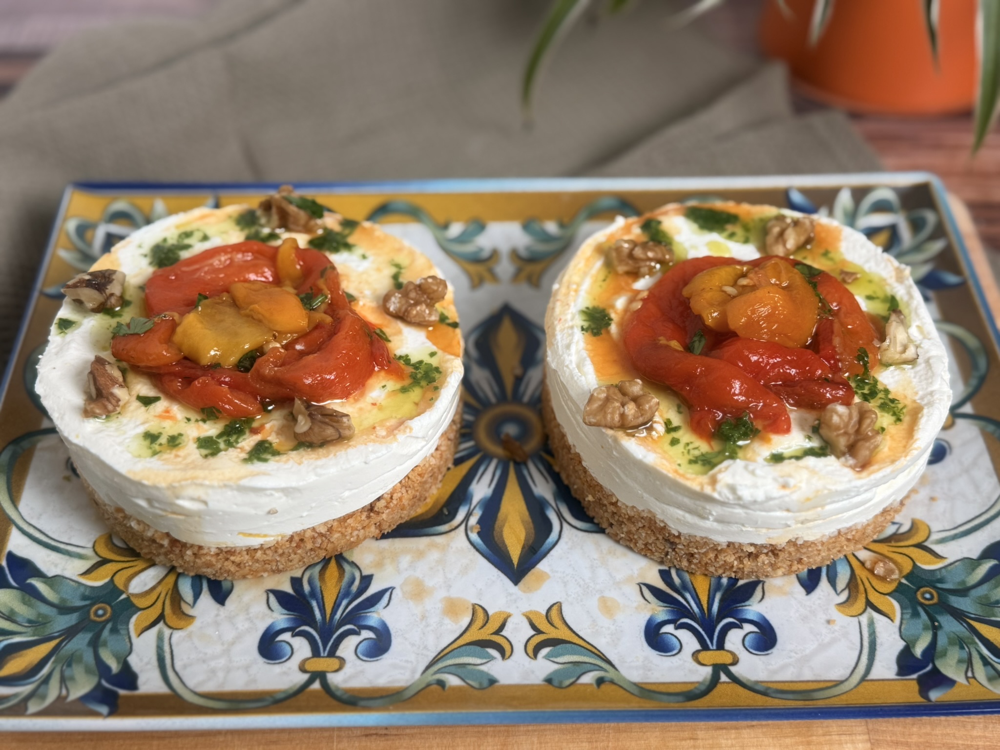 Mini cheesecake salate senza cottura: l’antipasto estivo facile e cremoso con base croccante di taralli, crema di formaggio spalmabile e robiola, topper di peperoni arrostiti, gherigli di noce e olio aromatizzato al prezzemolo, servite su un piatto bianco.