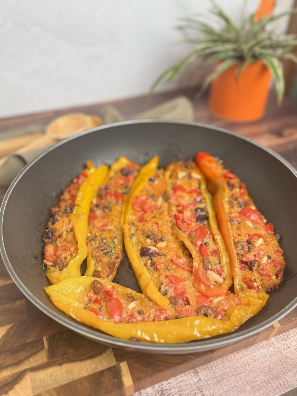 Peperoni ripieni in padella senza forno: facili, veloci e saporiti, ripieni di mollica di pane, pomodorini, parmigiano grattugiato, capperi, pinoli e basilico. Disposti su un piatto bianco. Il ripieno è dorato e leggermente gratinato, decorati con foglie di basilico fresco