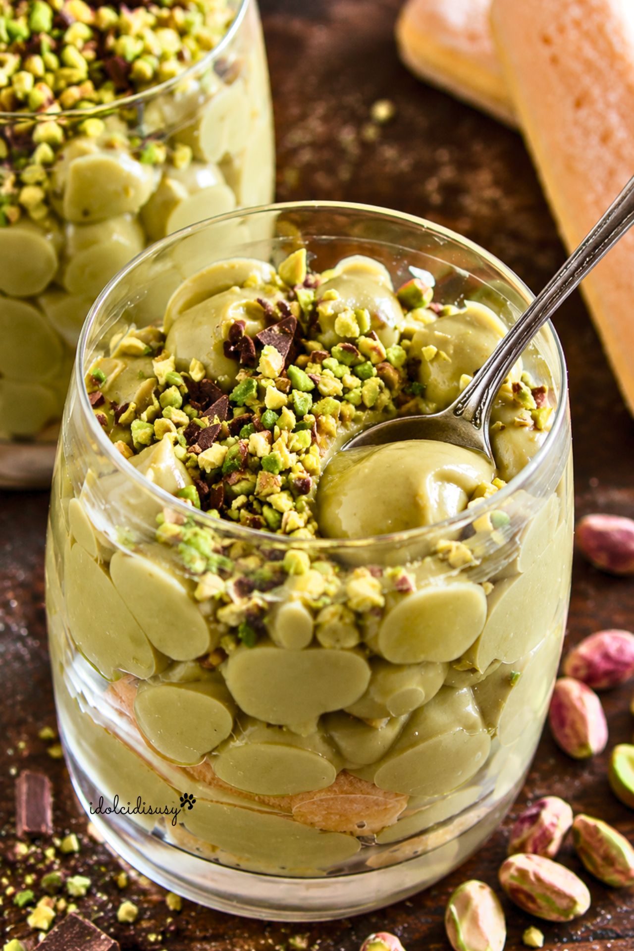 Dessert al cucchiaio con crema al pistacchio e savoiardi servito in bicchiere, decorato con pistacchi tritati e cioccolato fondente.