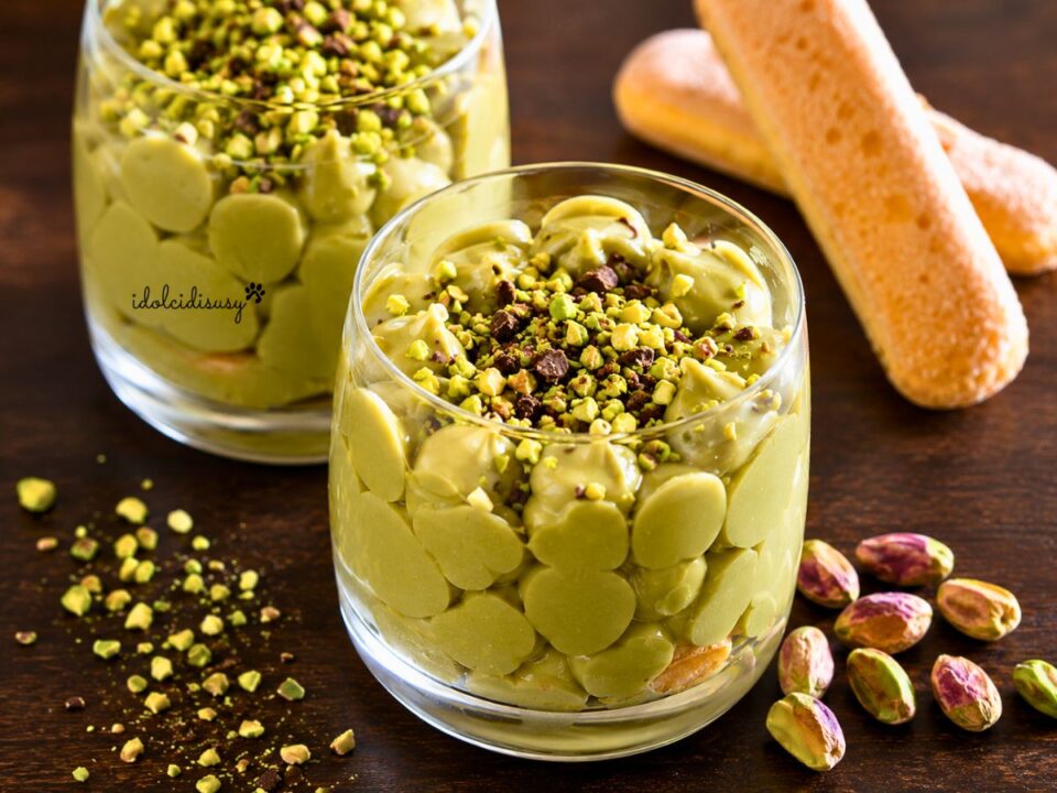 Due bicchieri di tiramisù al pistacchio con crema al pistacchio, savoiardi, granella di pistacchi e scaglie di cioccolato fondente su tavolo in legno.