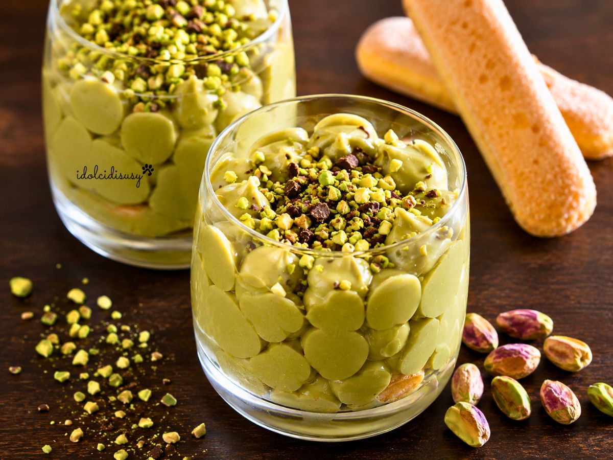 Due bicchieri di tiramisù al pistacchio con crema al pistacchio, savoiardi, granella di pistacchi e scaglie di cioccolato fondente su tavolo in legno.