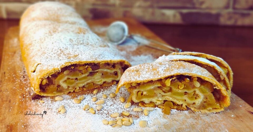 Strudel di mele dorato e spolverizzato con zucchero a velo, servito su tagliere di legno con pinoli e fetta tagliata