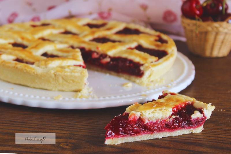Fetta di crostata con pasta brisée e ripieno di ciliegie succose, servita su piatto bianco con crostata intera sullo sfondo – idolcidisusy