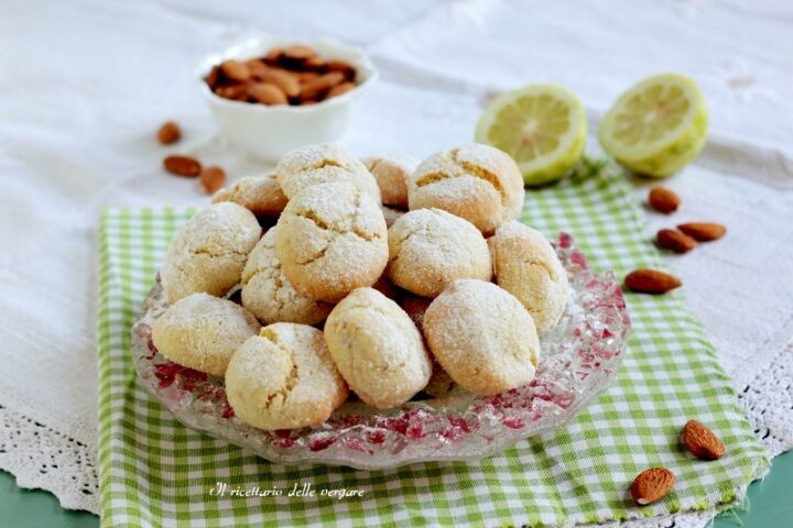 Biscotti morbidi al limone e mandorle ricoperti di zucchero a velo su un piatto di vetro con mandorle e limoni sullo sfondo.