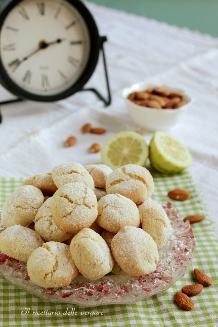 Biscotti morbidi al limone e mandorle in primo piano con un orologio vintage e una ciotola di mandorle sfocate sullo sfondo.