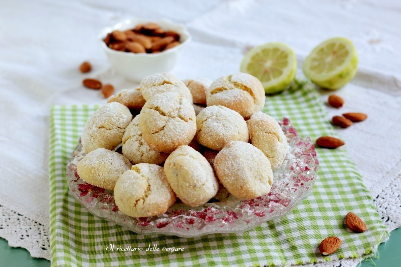 Biscotti morbidi al limone e mandorle ricoperti di zucchero a velo su un piatto di vetro con mandorle e limoni sullo sfondo.