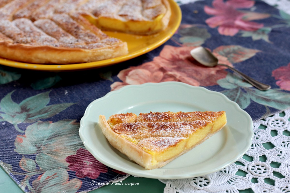Fetta di torta di pasta sfoglia e savoiardi su un piattino, con la torta intera sullo sfondo su una tovaglia floreale