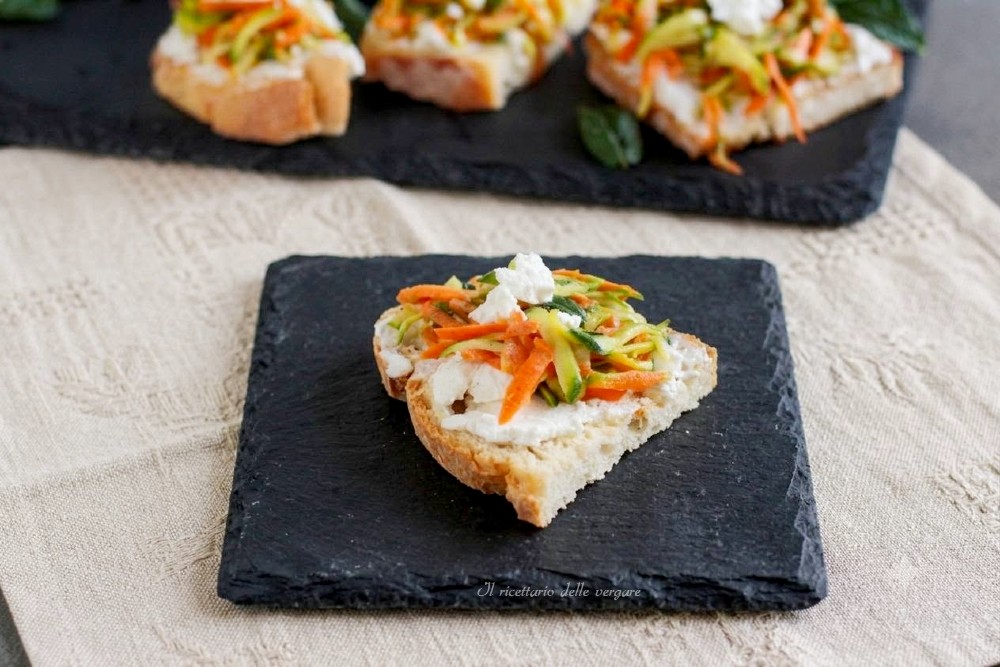 Bruschette con robiola di capra e julienne di zucchine e carote fresche servite su un piatto di pietra nera