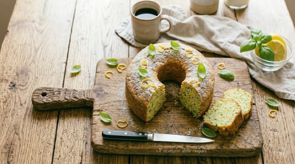 Ciambellone al basilico e limone tagliato a fette su un tagliere di legno rustico, decorato con zucchero a velo, zeste di limone e foglie di basilico fresco.