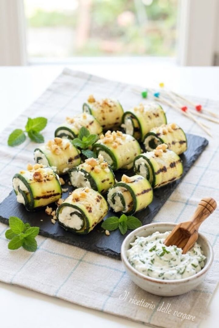 Ciotola di robiola condita con erbe aromatiche accanto a vassoio di involtini di zucchine grigliate con noci e menta.